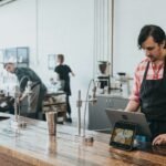A barista in a plaid shirt and apron using a tablet at a wooden counter