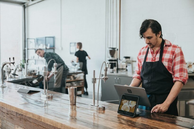 A barista in a plaid shirt and apron using a tablet at a wooden counter